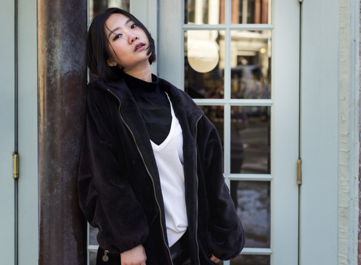 Young Asian woman in a black turtleneck and white top under an oversized fluffy fleece jacket, posing outside a blue storefront, showcasing Korean tops and layered streetwear.