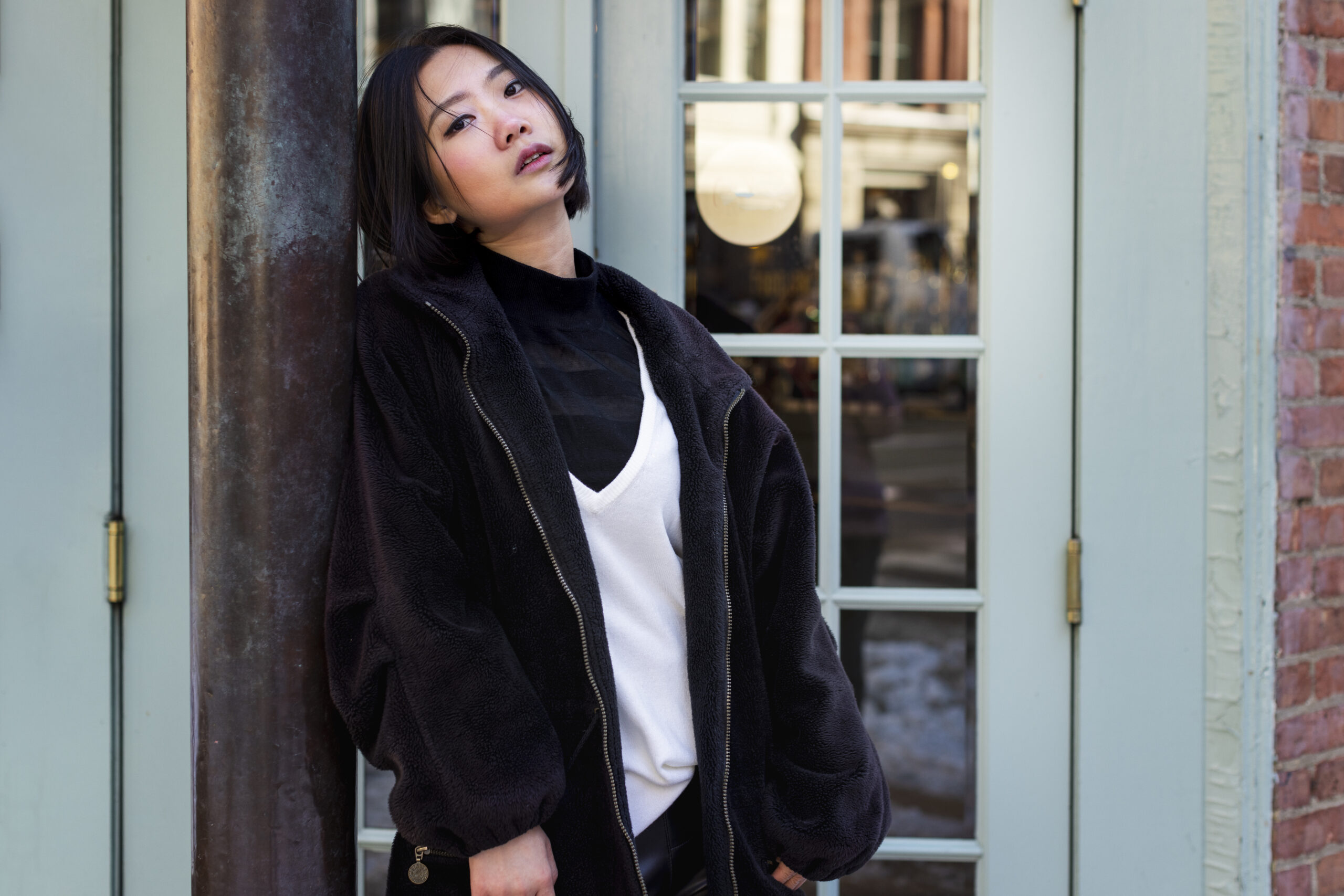 Young Asian woman in a black turtleneck and white top under an oversized fluffy fleece jacket, posing outside a blue storefront, showcasing Korean tops and layered streetwear.